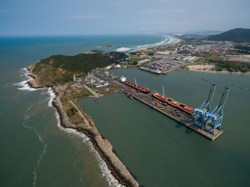 Vista aérea do Porto de Imbituba, no Brasil, com cais, guindastes, embarcações e trecho da costa.