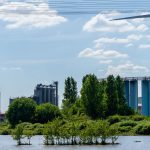 Paisagem industrial com fábricas, uma turbina eólica e vegetação às margens de um corpo d’água.