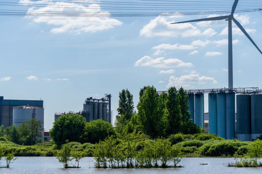 Paisagem industrial com fábricas, uma turbina eólica e vegetação às margens de um corpo d’água.
