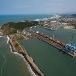 Vista aérea do porto de Imbituba, com navios, guindastes, docas e a costa brasileira.