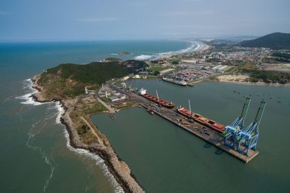 Vista aérea do porto de Imbituba, com navios, guindastes, docas e a costa brasileira.