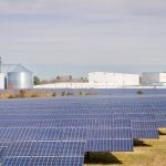 Campo de painéis solares diante de galpões e silos industriais sob céu claro.